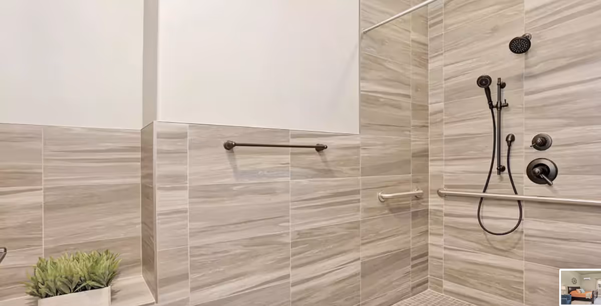 Modern tiled walk-in shower with a handheld showerhead, horizontal grab bars and a small potted plant on a ledge.