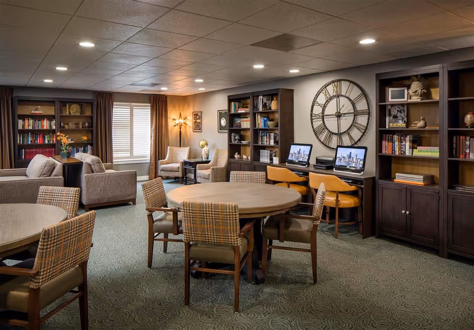 A cozy senior living common area with round wooden tables surrounded by plaid upholstered chairs. There are two beige armchairs near a window with brown curtains, and two sofas with a small table between them. The room features dark wooden bookshelves filled with books and decorative items, a large wall clock, and a desk area with two computers and orange chairs. The carpet is green with a subtle pattern, and the ceiling has recessed lighting.