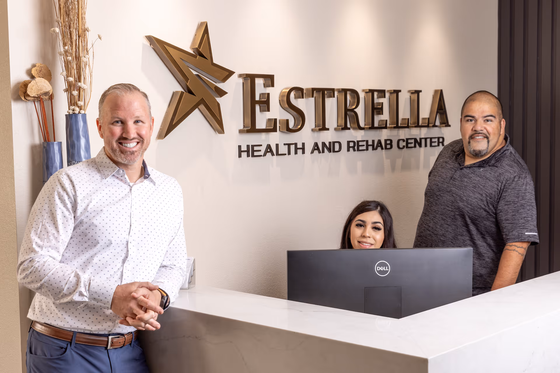Three staff members at the reception desk of Estrella Health and Rehab Center. One man in a white shirt stands smiling on the left, a woman sits behind a computer monitor in the center, and another man in a dark shirt stands on the right. The facility's name and logo are displayed on the wall behind them.