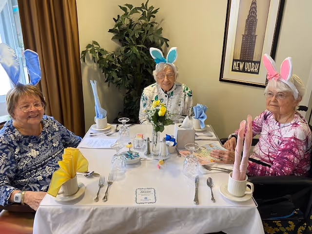 Three elderly women sitting around a dining table set with plates, glasses, and folded napkins. Two of the women are wearing bunny ear headbands, one blue and one pink. There is a vase with yellow and white flowers in the center of the table, and a framed New York poster on the wall behind them.