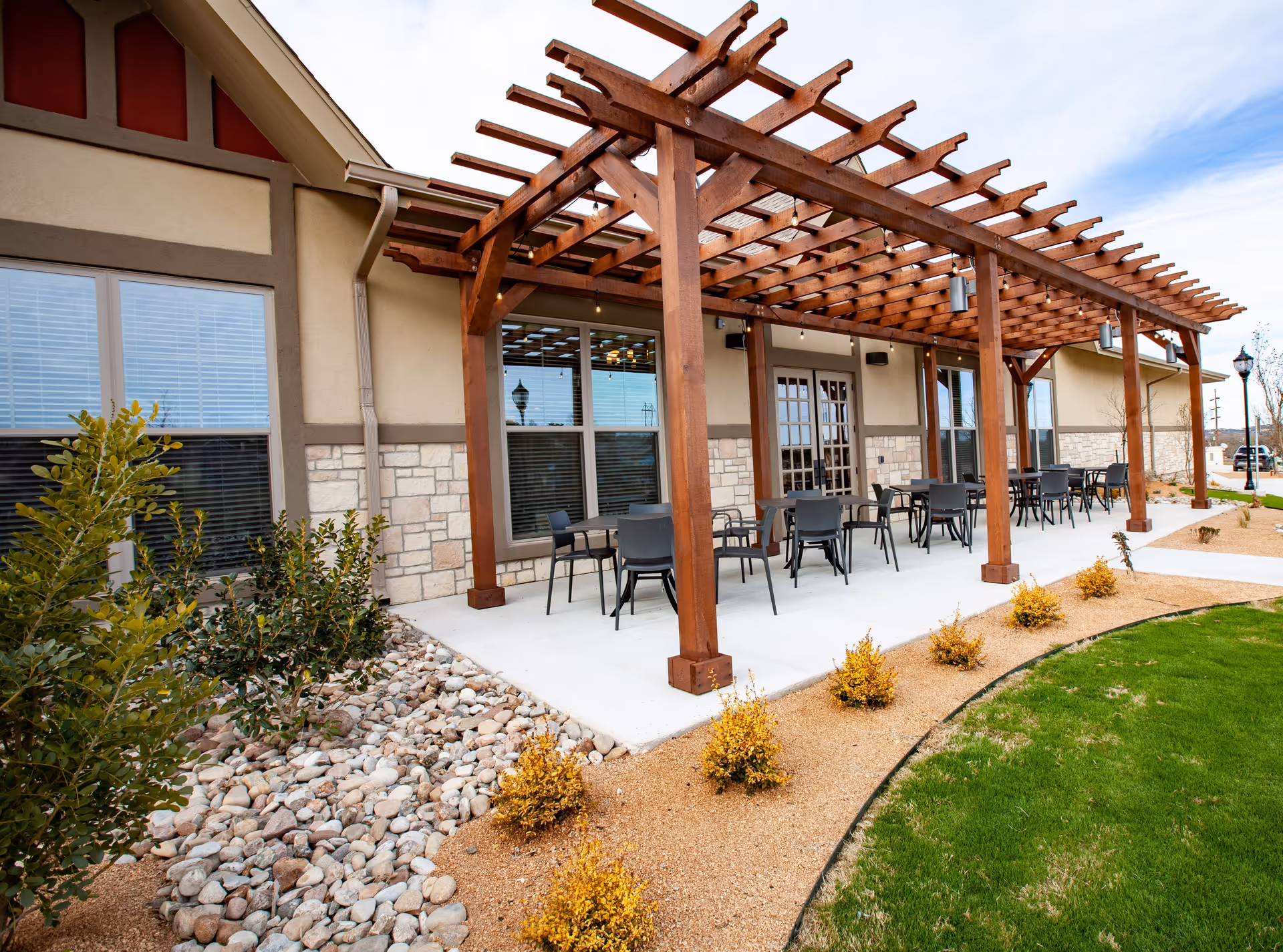 Outdoor patio area at The Villages of Windcrest featuring a wooden pergola with string lights, several tables and chairs arranged on a concrete floor, surrounded by landscaped plants, rocks, and green grass under a partly cloudy sky.