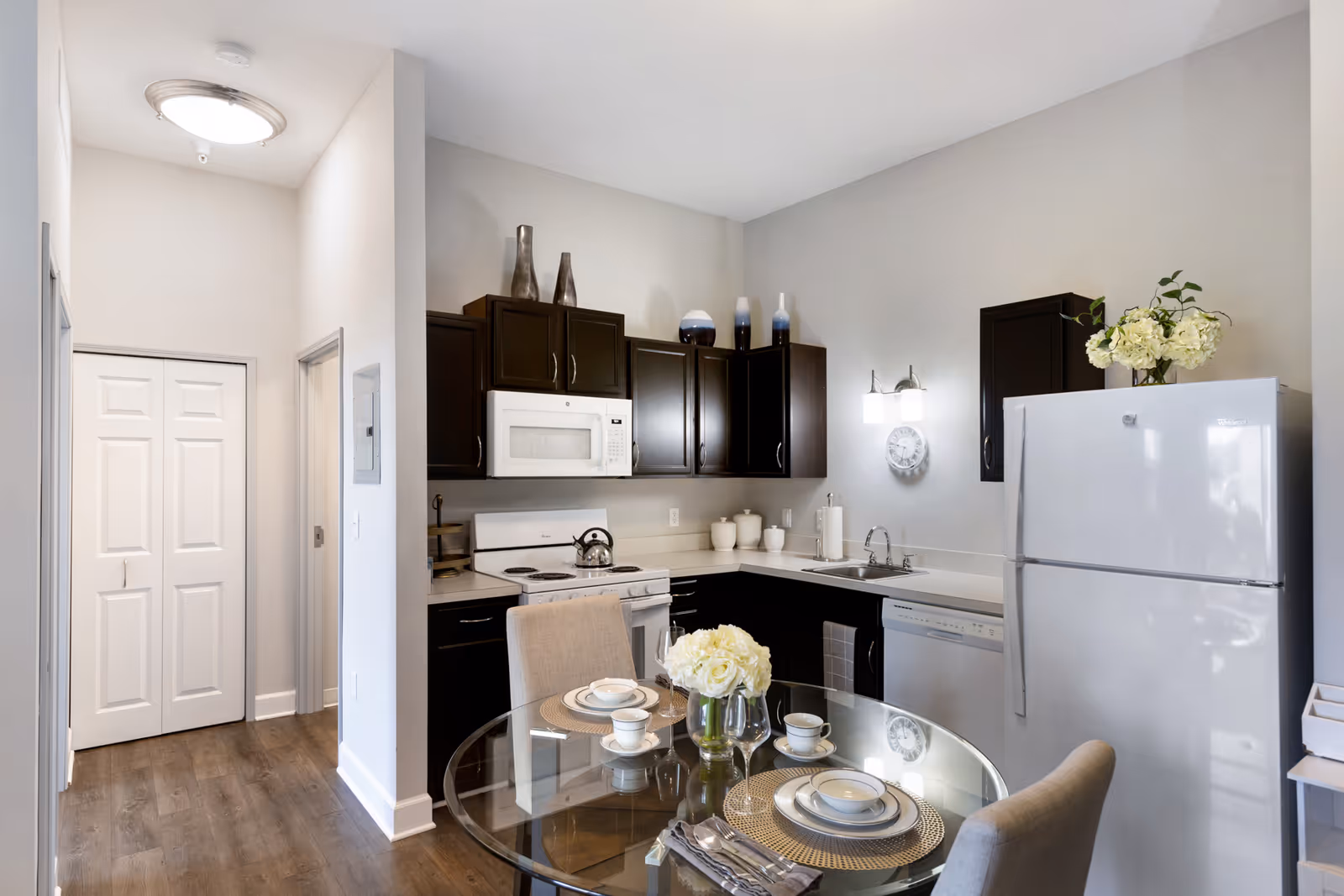 Small modern kitchen and dining area with dark cabinets, white appliances, and a glass-top table set for two.