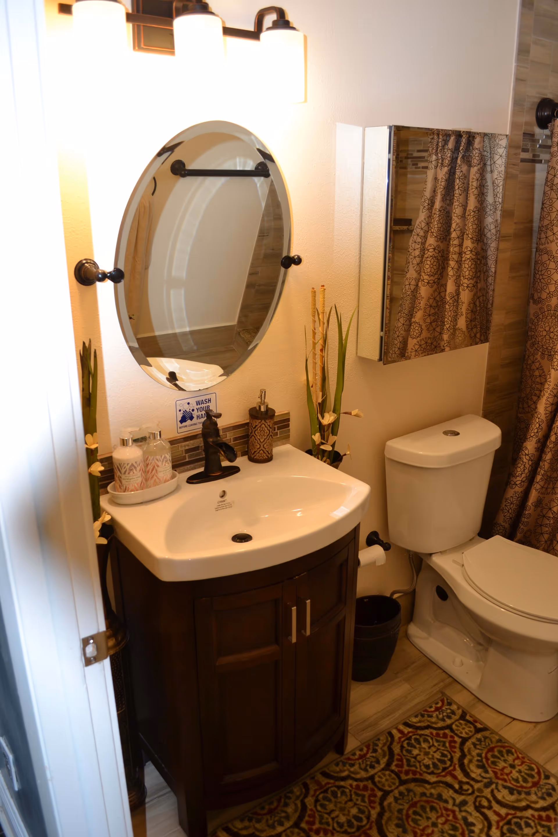 A small bathroom with a white sink set in a dark wooden vanity, an oval mirror above the sink, a toilet next to the vanity, a patterned shower curtain, and a decorative rug on the floor. The sink area has soap dispensers and decorative plants on either side. A wall-mounted cabinet with a mirrored door is above the toilet.
