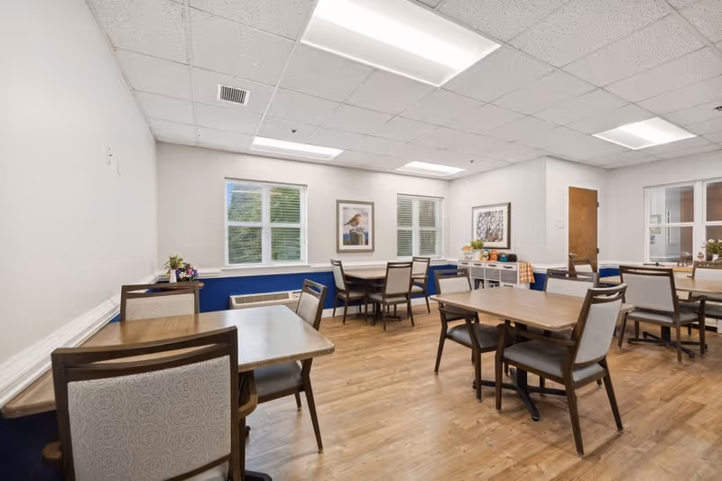 A bright dining room with several wooden tables and cushioned chairs arranged neatly. The room has light-colored walls with a blue accent on the lower half, wood flooring, and a drop ceiling with fluorescent lights. There are two windows with white blinds, framed artwork on the walls, and a sideboard with decorative items and a checkered cloth near a wooden door.