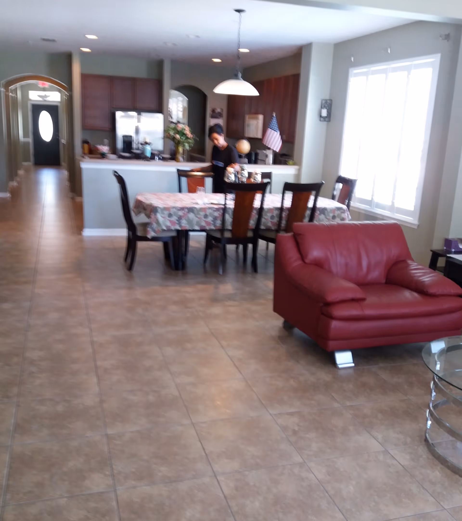 Interior view of a living and dining area with tiled flooring. A red leather armchair is positioned near a glass coffee table. A dining table covered with a floral tablecloth is surrounded by six chairs. A person is standing near the kitchen counter in the background, which has wooden cabinets, a refrigerator, and an American flag. Large windows with white shutters allow natural light into the room.