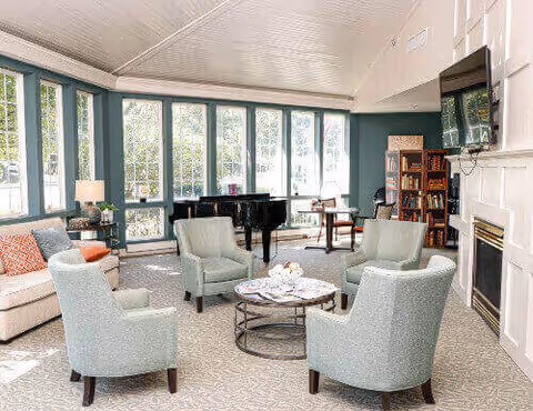 A bright and spacious living room with large windows letting in natural light. The room features a white sofa with orange and patterned cushions, four light blue upholstered armchairs arranged around a round coffee table with magazines, a black grand piano near the windows, a fireplace with a mounted TV above it, and a bookshelf filled with books and decorative items.