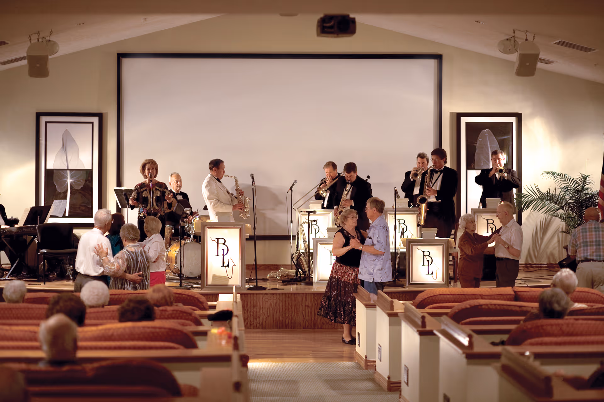 A live band performs on stage in a community hall while several elderly couples dance in front of the stage. The audience is seated in rows of cushioned chairs facing the stage. The room is warmly lit and decorated with framed artwork and plants.