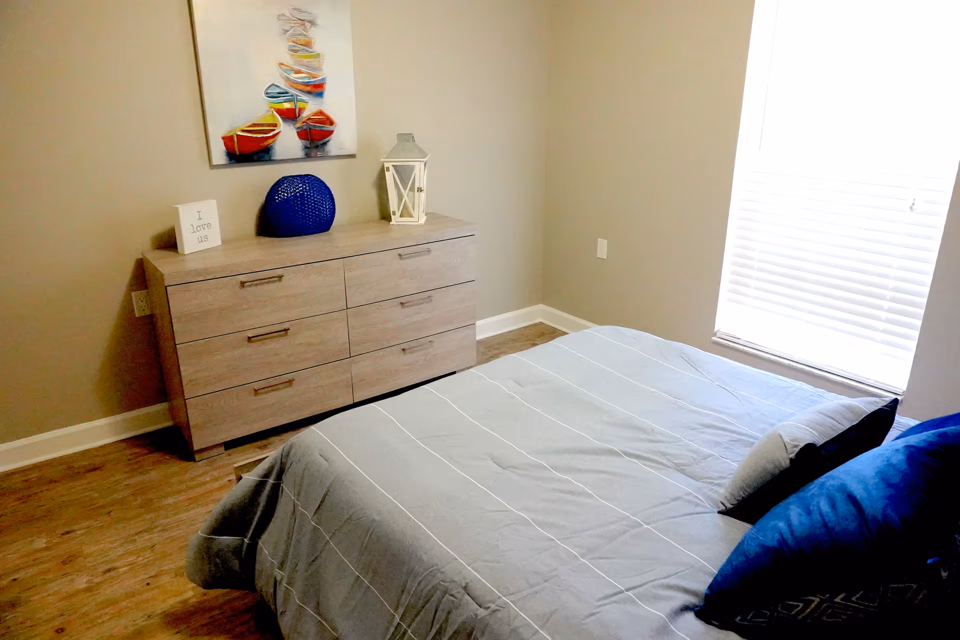 A bedroom with a bed covered in a gray bedspread and blue pillows. Next to the bed is a wooden dresser with six drawers, decorated with a blue vase, a white lantern, and a small sign that says 'I love us'. Above the dresser hangs a colorful painting of boats. A window with white blinds is on the right side of the room.
