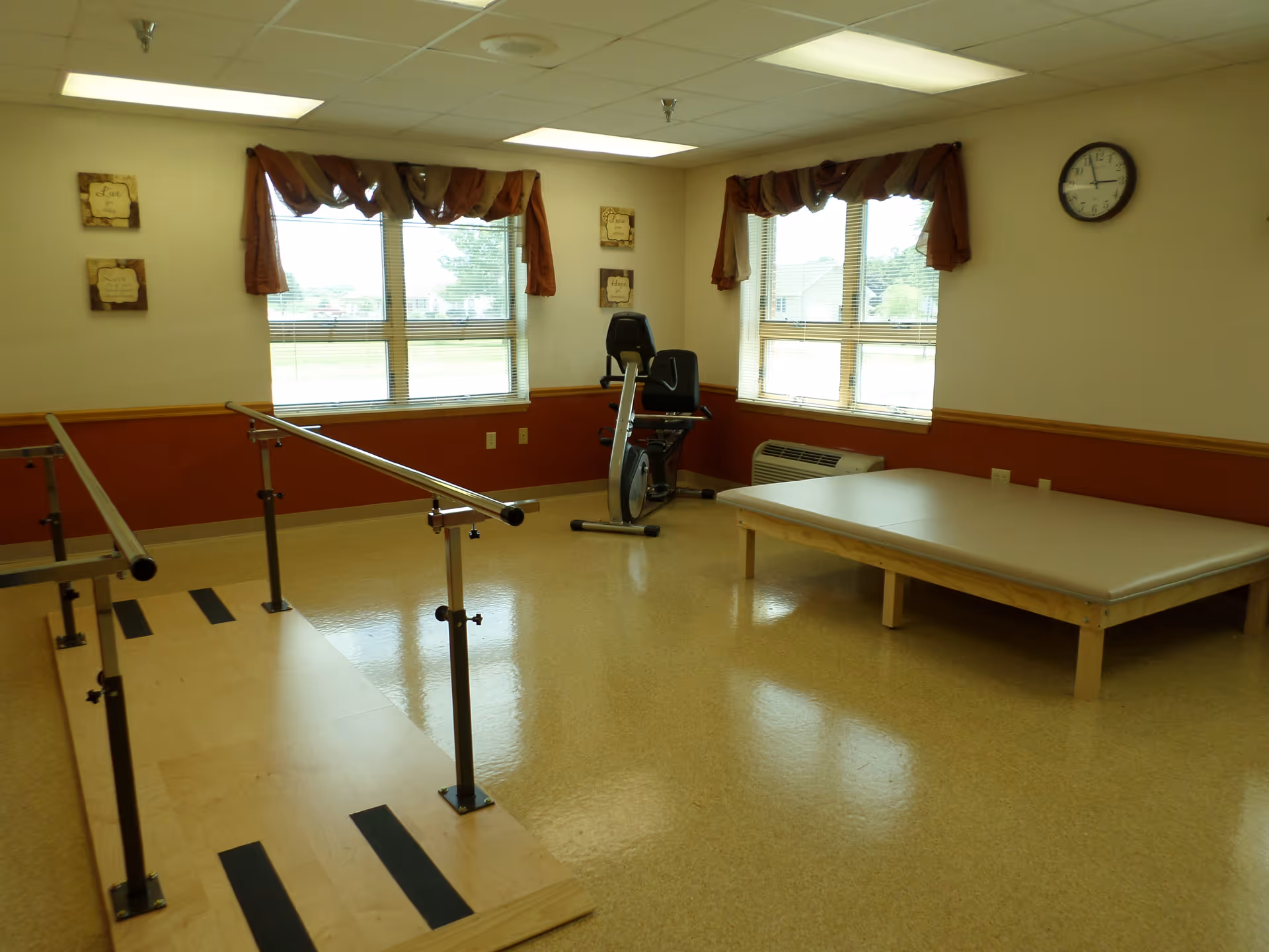 A rehabilitation room with parallel bars for walking exercises, a padded therapy table, and a stationary exercise bike. The room has two large windows with brown valances, a wall clock, and motivational wall plaques.