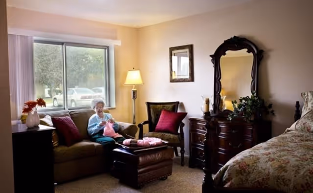 A cozy bedroom in a senior living facility with a large window showing parked cars outside. An elderly woman is sitting on a brown sofa knitting with pink yarn. The room features a floral bedspread on a wooden bed, a wooden dresser with an ornate mirror, a side table with a vase of flowers, a floor lamp, and a cushioned armchair with a red pillow.