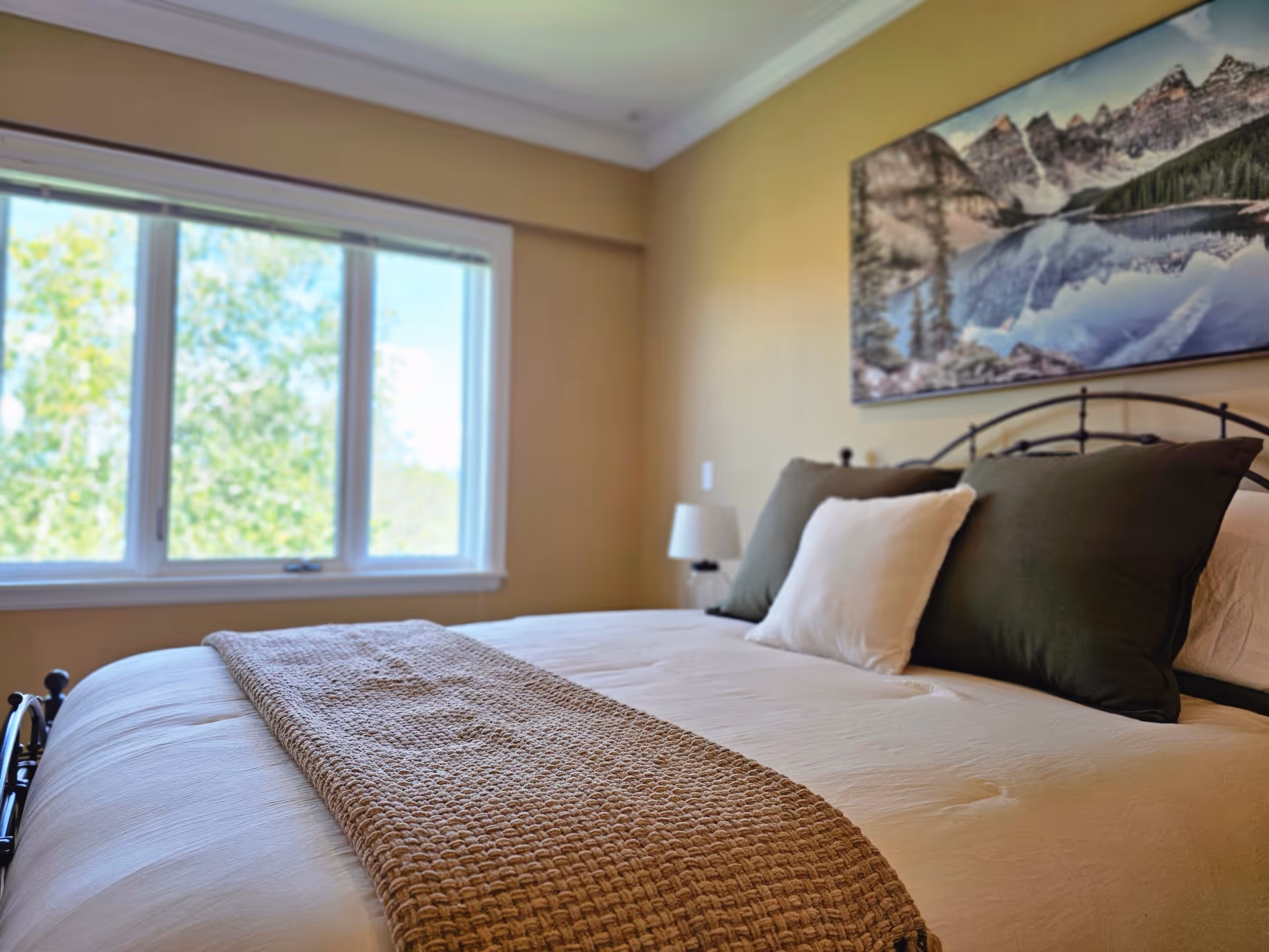 Bright bedroom with a neatly made bed topped with green and white pillows, a textured runner, a window letting in daylight, and landscape artwork above the headboard.