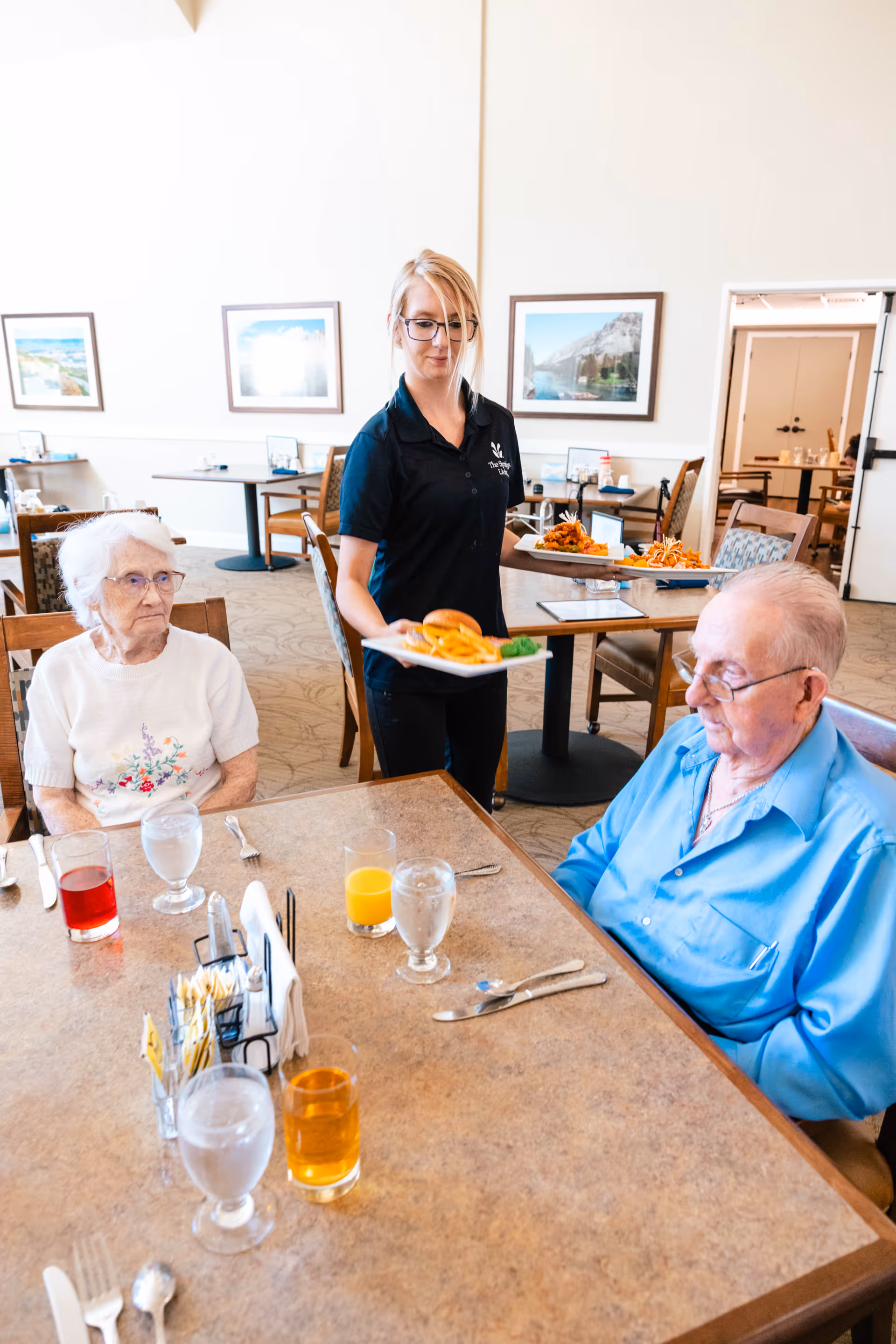 A caregiver serves plates of food to two elderly residents seated at a dining table in a bright dining room.