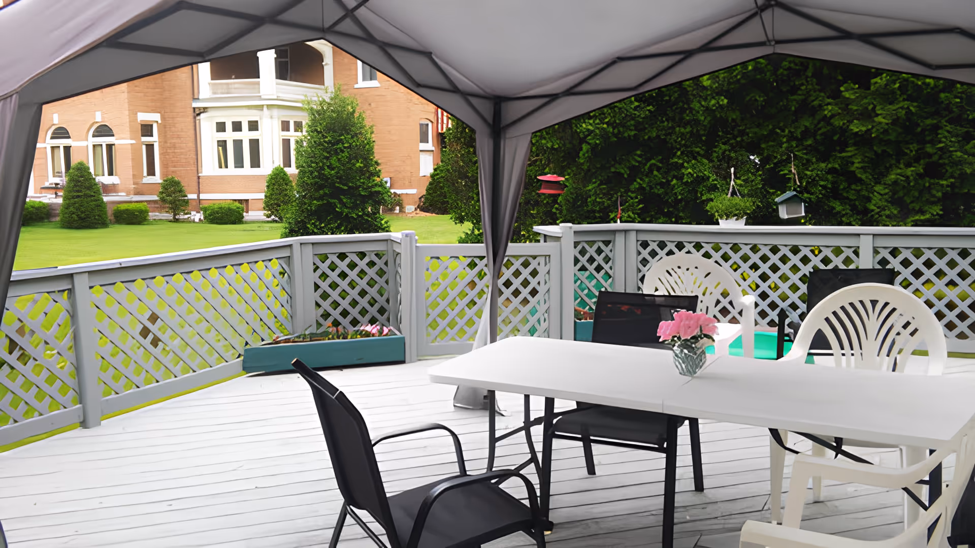 Outdoor patio area with a white table and mixed black and white chairs under a canopy. The patio has a white lattice railing and overlooks a green lawn with trees and a brick building in the background. A small vase with pink flowers is on the table.