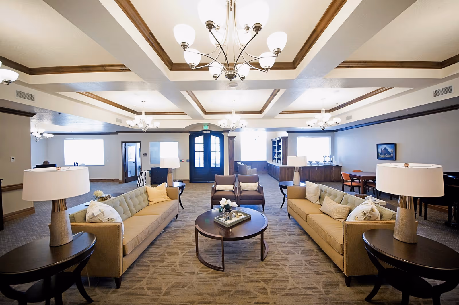 Spacious furnished common lounge with sofas, armchairs, coffee table, lamps, and chandeliers in an assisted living facility.