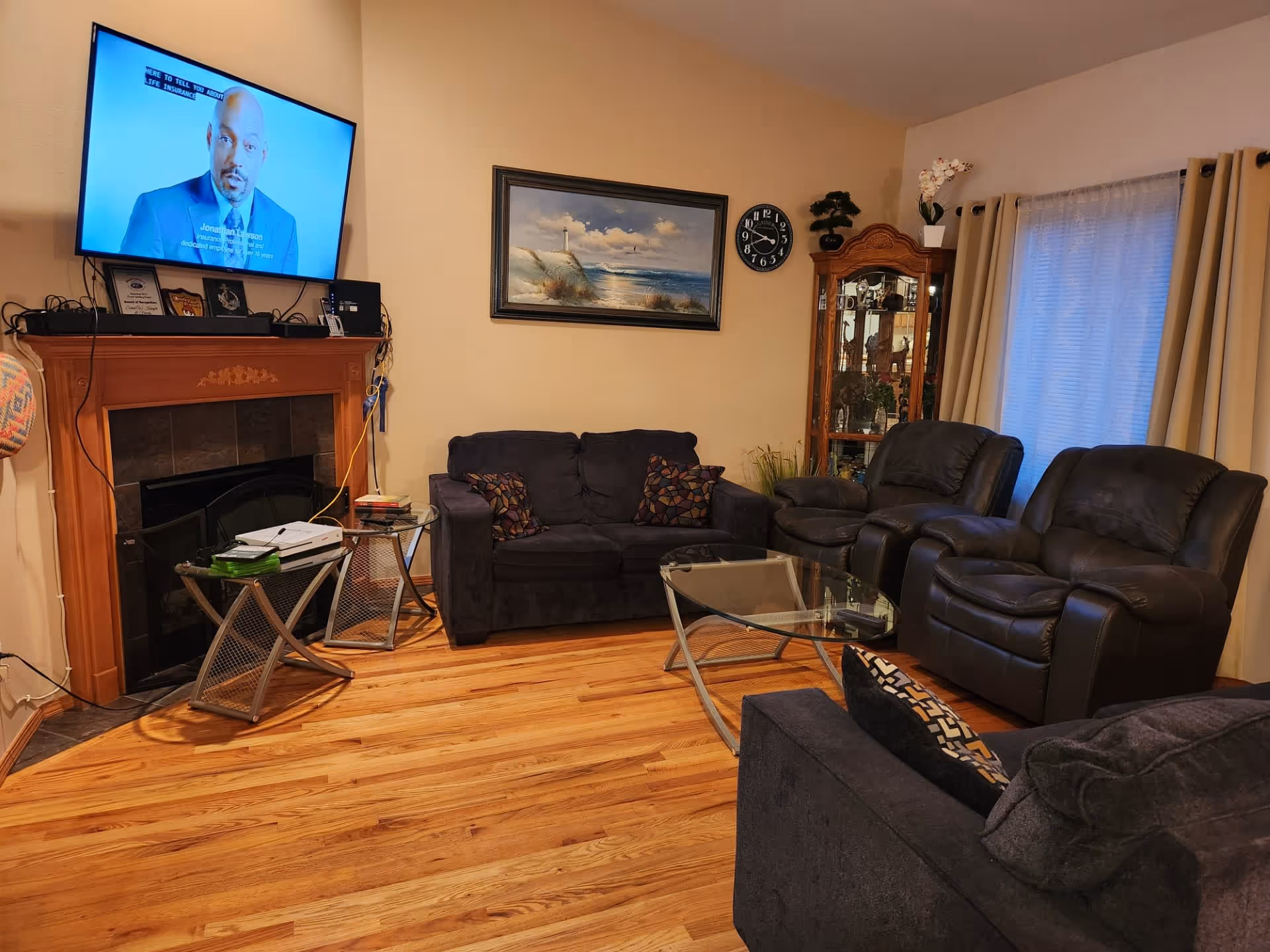 A cozy living room with wooden flooring, featuring a dark blue loveseat and matching sofa with patterned cushions, two black leather recliners, a glass coffee table, and a wooden fireplace with a mounted flat-screen TV above it. A framed painting of a lighthouse and ocean hangs on the wall, with a round clock nearby. A glass display cabinet with decorative items is positioned near a window with beige curtains.