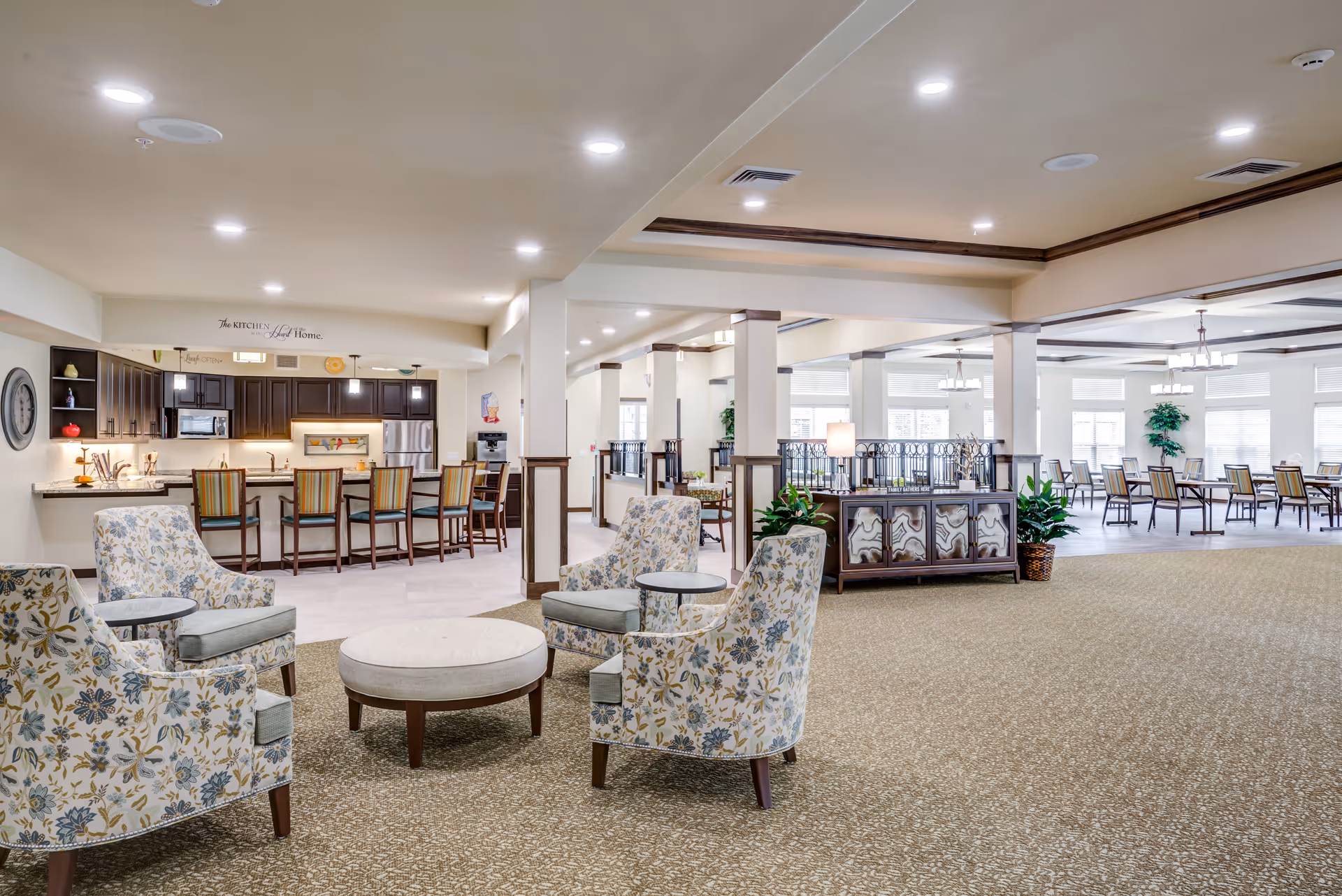 Bright senior living common area with upholstered armchairs around a round table, a bar with stools and kitchenette at the back and dining tables in the background.
