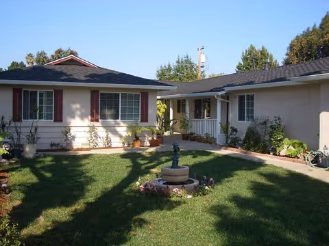 A sunny courtyard area with green grass and a small fountain surrounded by flowers in the center. The courtyard is enclosed by single-story buildings with windows, plants, and a small porch area. Trees and a clear blue sky are visible in the background.
