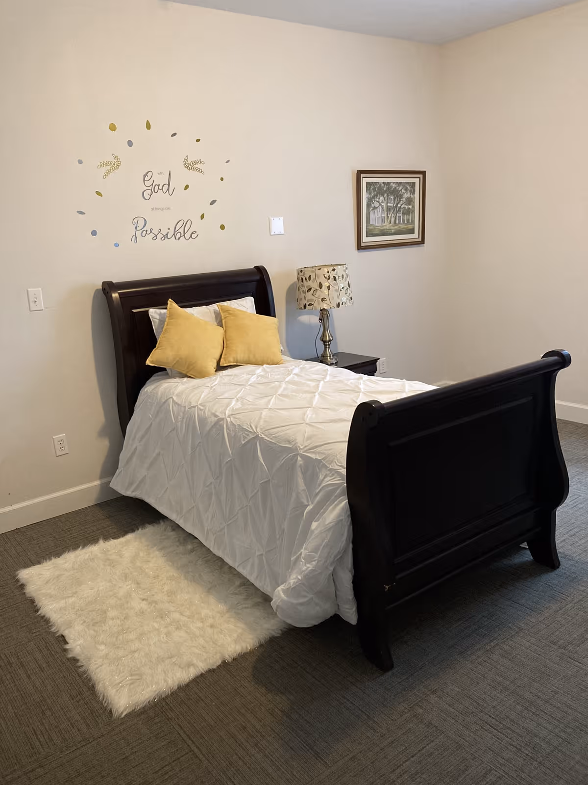 A bedroom with a dark wooden bed frame, white bedding, and two yellow pillows. There is a small nightstand with a decorative lamp next to the bed. On the wall above the bed is a decal that reads 'God makes all things Possible' surrounded by small decorative leaves. A framed picture hangs on the adjacent wall, and a small white fluffy rug is placed on the floor beside the bed.