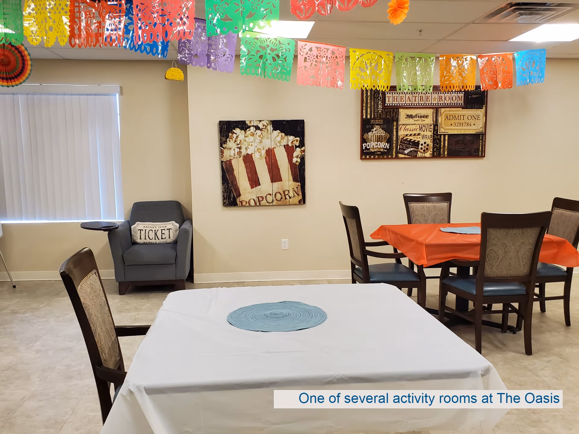 Activity room decorated with colorful paper banners hanging from the ceiling, tables covered with white and orange tablecloths, chairs around the tables, a gray armchair with a pillow that says 'Admit One Ticket', and wall art featuring popcorn and movie themes.