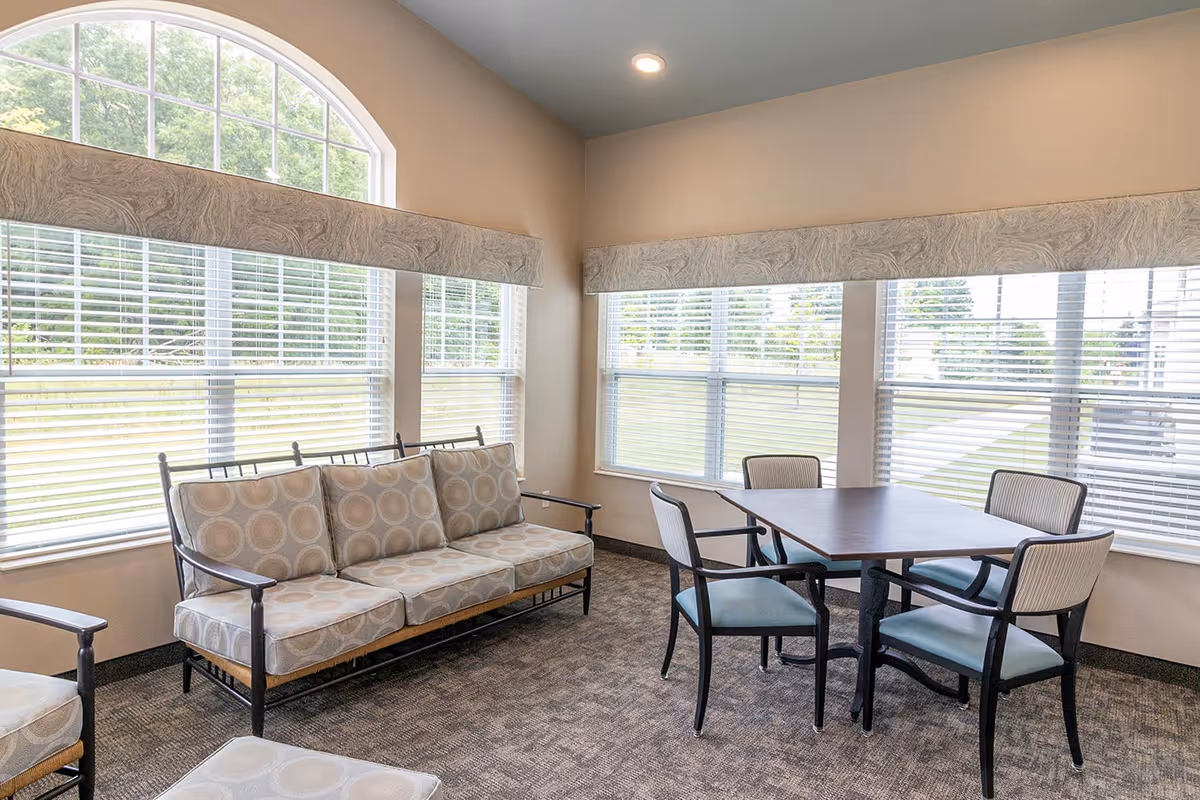 A bright room with large windows featuring white blinds and valances. The room contains a cushioned sofa with patterned upholstery and a square table surrounded by four chairs with light blue seats. The carpet is patterned in neutral tones, and the walls are painted light beige.