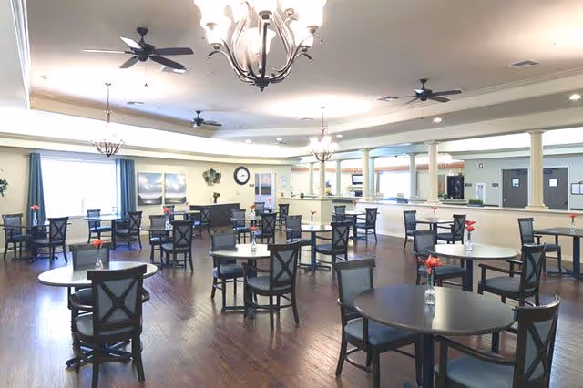 A spacious dining room with multiple round tables and chairs arranged neatly. The room has wooden flooring, ceiling fans, chandeliers, and large windows with curtains allowing natural light to enter. Small vases with flowers are placed on each table.
