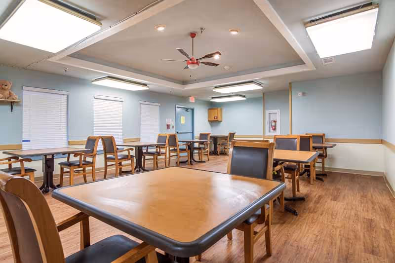 A bright dining room with multiple wooden tables and chairs arranged neatly on a wood floor. The walls are painted light blue with white trim, and there are three windows with closed blinds. The ceiling has recessed lighting, a ceiling fan with red blades, and two skylights. A teddy bear sits on a small shelf on the left wall.