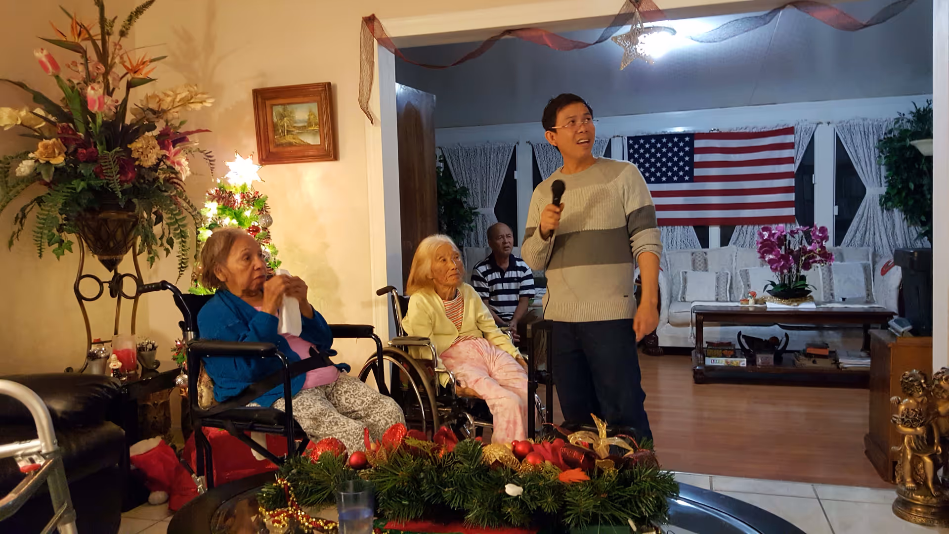 A man holding a microphone stands in a living room decorated for Christmas, with two elderly women in wheelchairs and an elderly man seated nearby. The room features a Christmas tree with lights, an American flag hanging on the wall, floral arrangements, and comfortable seating.