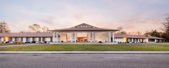Wide exterior view of a single-story building with a large covered entrance supported by columns, surrounded by a well-maintained lawn and landscaping, under a soft evening sky.