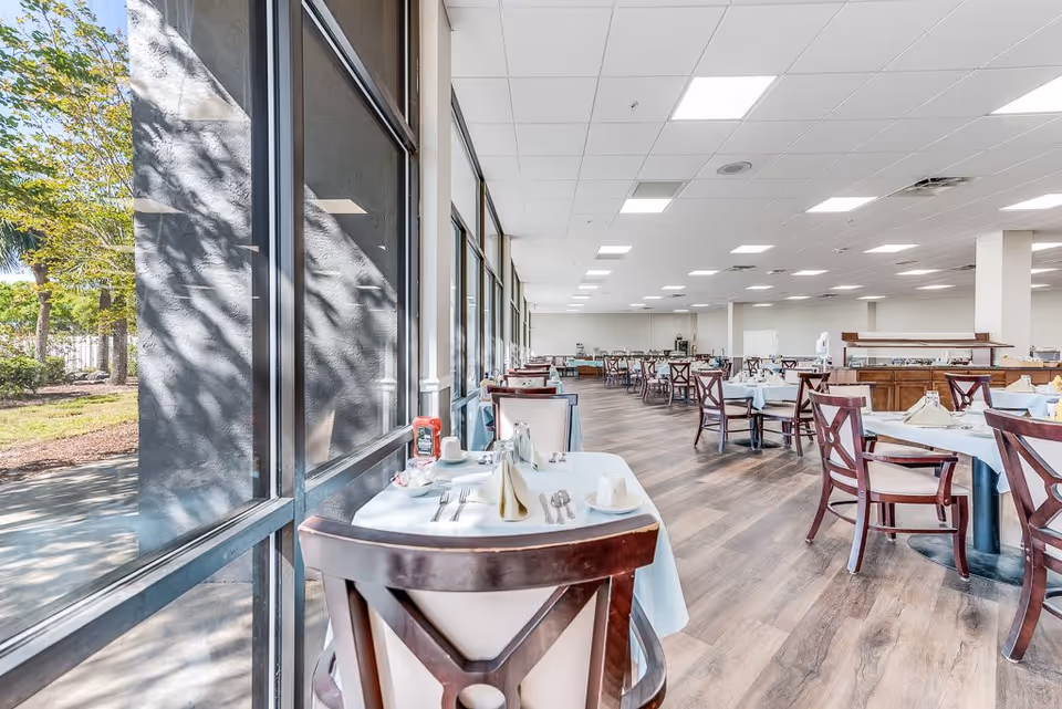 Bright and spacious dining room with large windows on the left side letting in natural light. Multiple tables are set with white tablecloths, napkins, utensils, and condiments. Wooden chairs with cushioned seats surround the tables. The floor is wood-patterned, and the ceiling has recessed lighting. Outside greenery is visible through the windows.