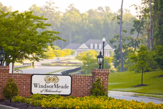 Entrance sign for WindsorMeade of Williamsburg set in a brick structure with two black lanterns on either side, surrounded by green trees and yellow flowers, with a large house and wooded area in the background.