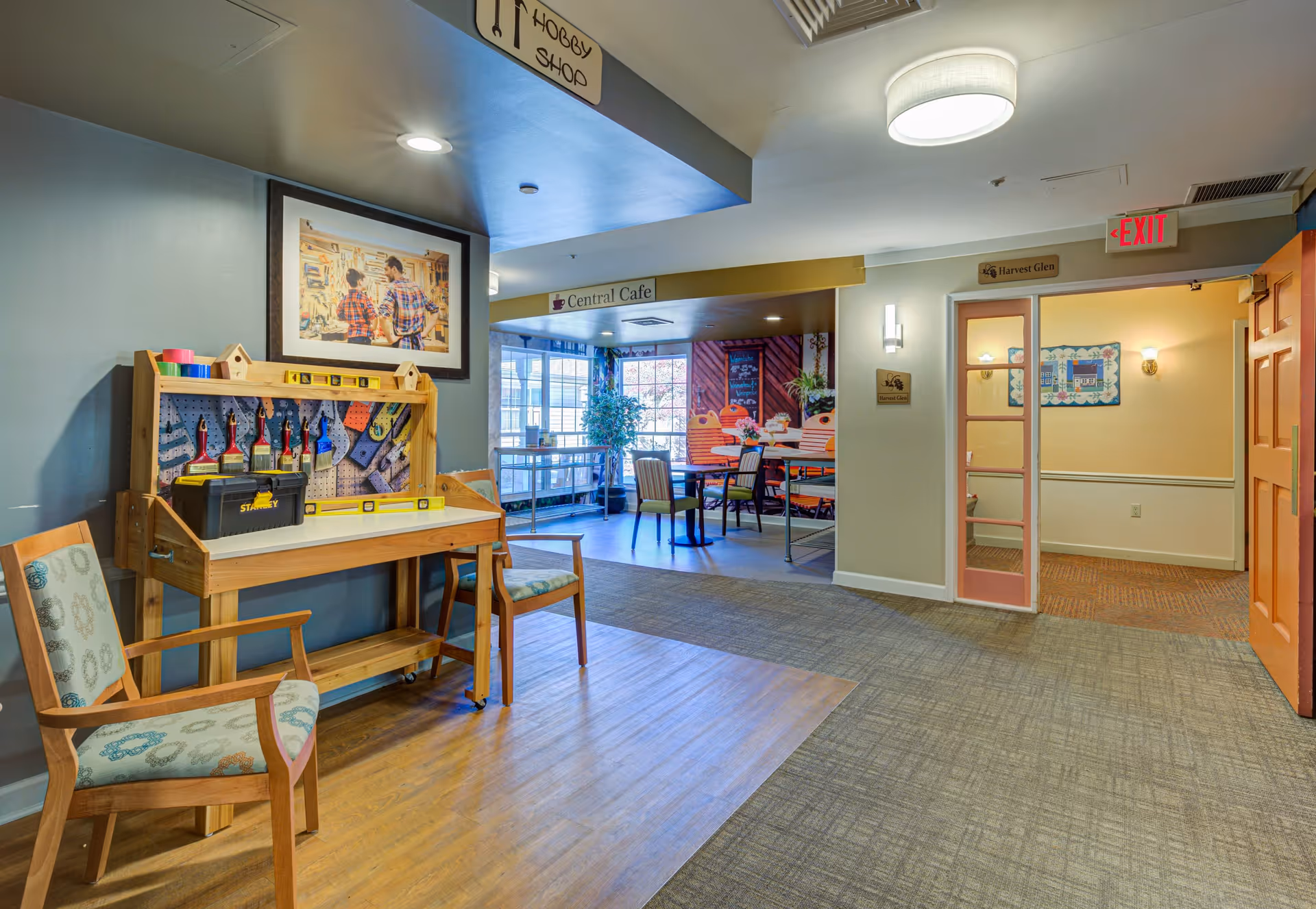 Interior view of a senior living facility hallway featuring a hobby shop area with a wooden workbench, tools, and two chairs. In the background, there is a dining area labeled Central Cafe with tables and chairs near large windows letting in natural light. The hallway has carpet and wood flooring, overhead lighting, and signs indicating different areas including Harvest Glen and an exit door.