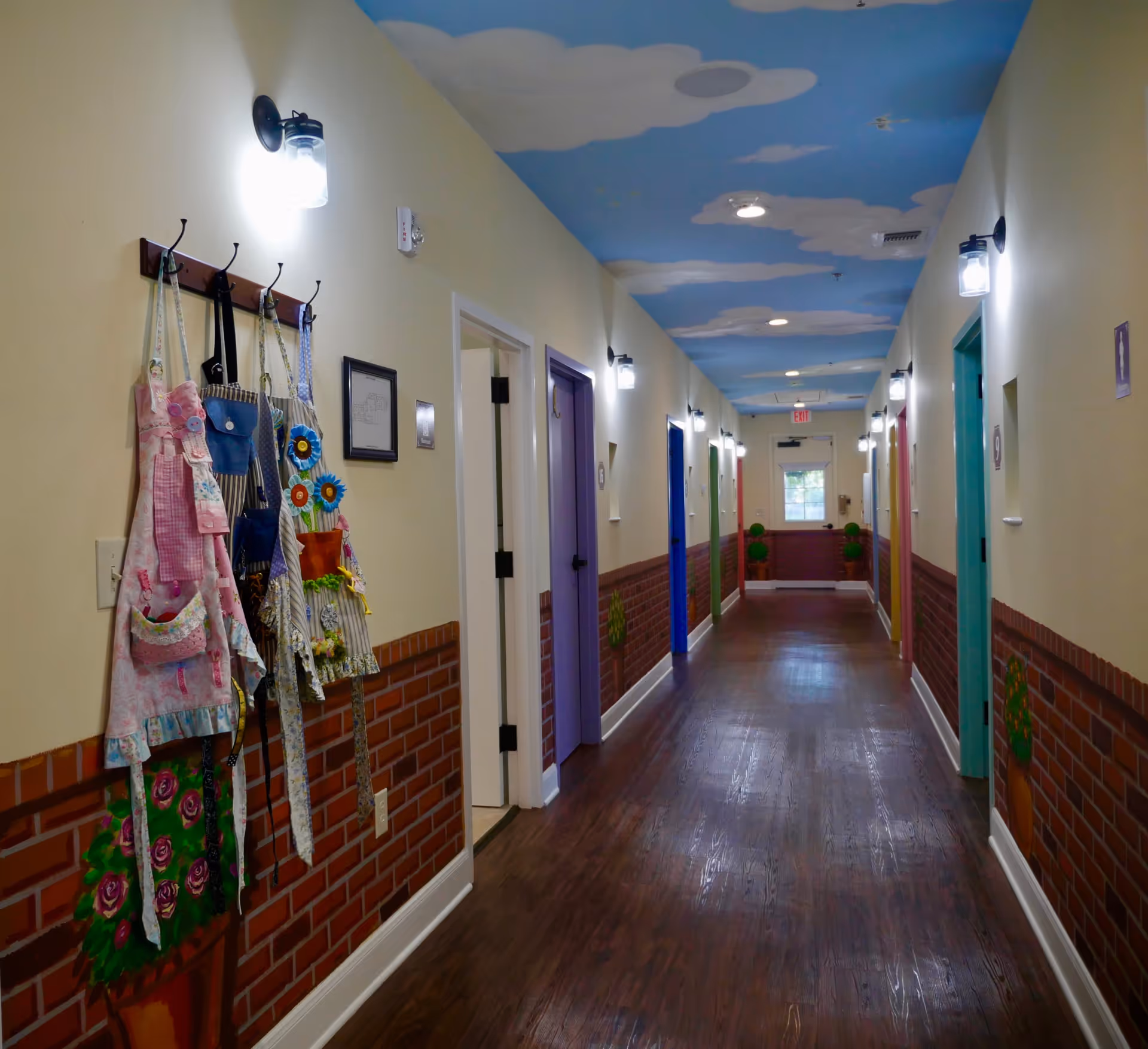 A brightly lit hallway with wooden flooring and walls painted to resemble a brick lower half and cream upper half. Several colorful doors line the hallway, each with a number plaque. The ceiling is painted blue with white clouds, creating a cheerful atmosphere. Aprons hang on hooks on the left wall near the entrance, and there are small decorative flower paintings along the walls.