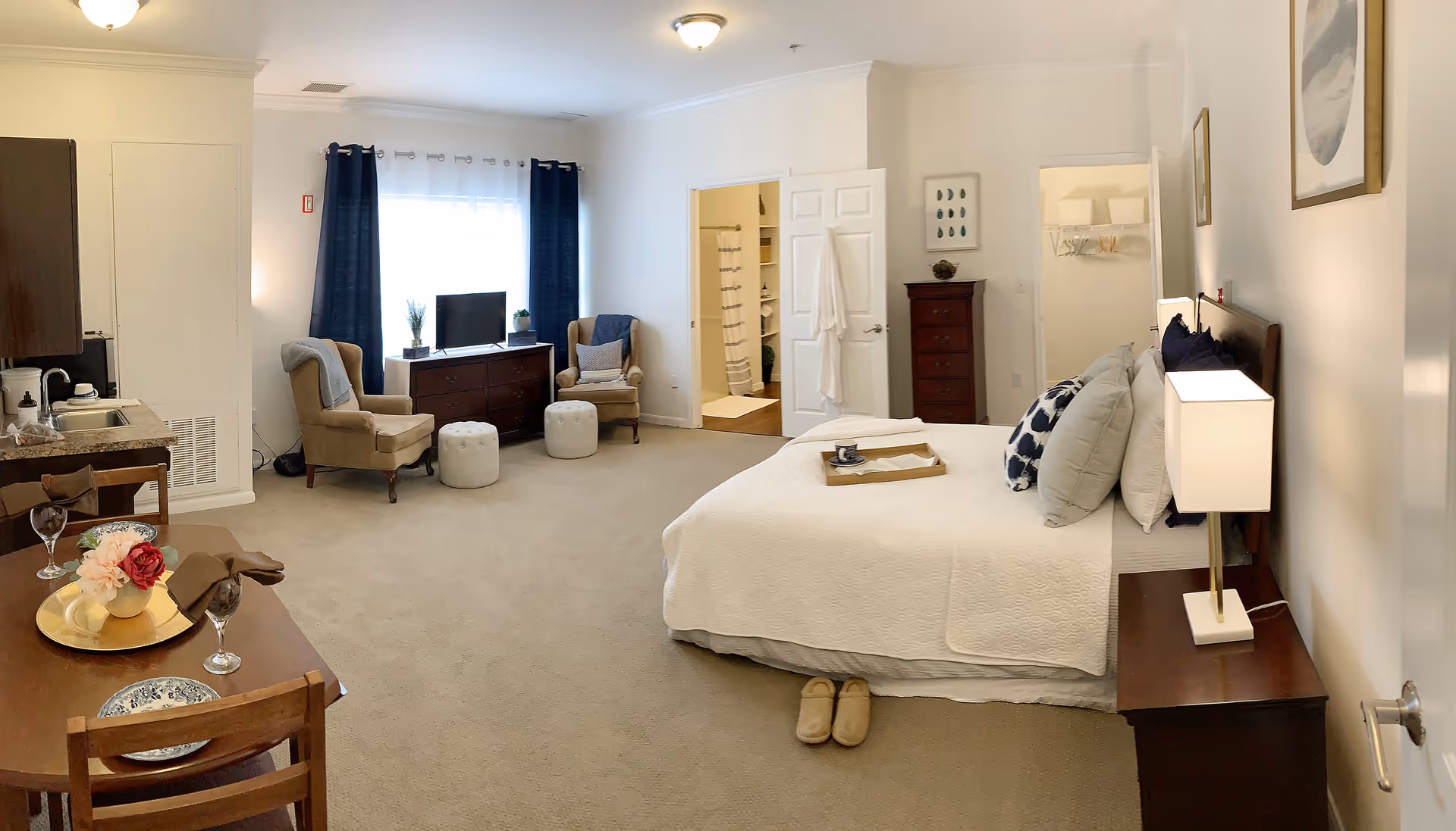 A spacious senior living bedroom with a large bed covered in white bedding and multiple pillows. There is a wooden nightstand with a lamp beside the bed. At the foot of the bed, a pair of slippers is placed on the carpeted floor. The room also features a small dining table set for two with plates, glasses, and a floral centerpiece. Two beige armchairs with a small ottoman are positioned near a window with dark blue curtains. A dresser with a TV and decorative plants is against the wall. Two open doors reveal a bathroom with a shower curtain and a walk-in closet with shelves and hangers. The walls are painted white, and the room is well-lit with ceiling lights.