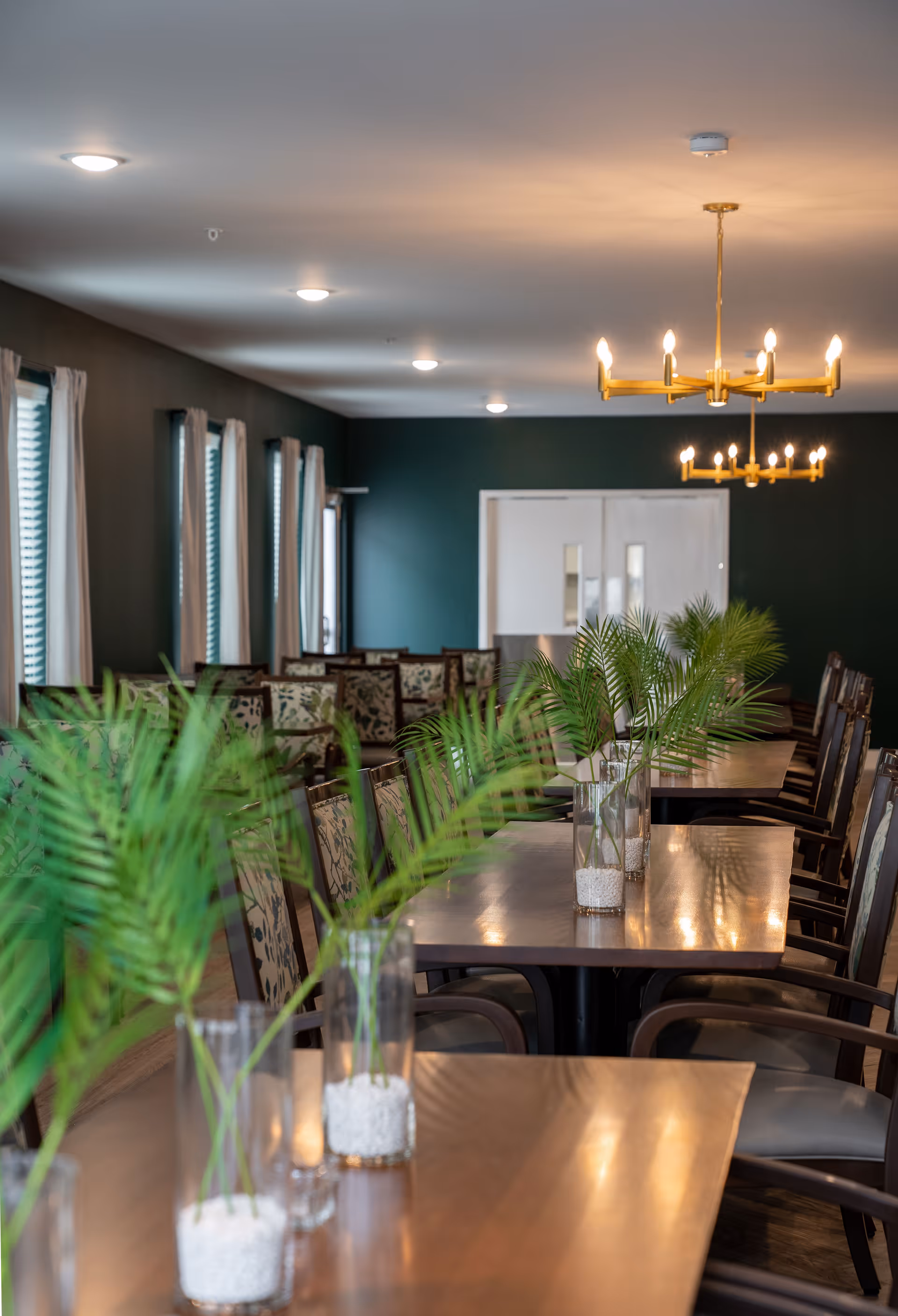 Long communal dining room with wooden tables and chairs, vases of palm fronds as centerpieces, chandeliers, and windows with curtains.