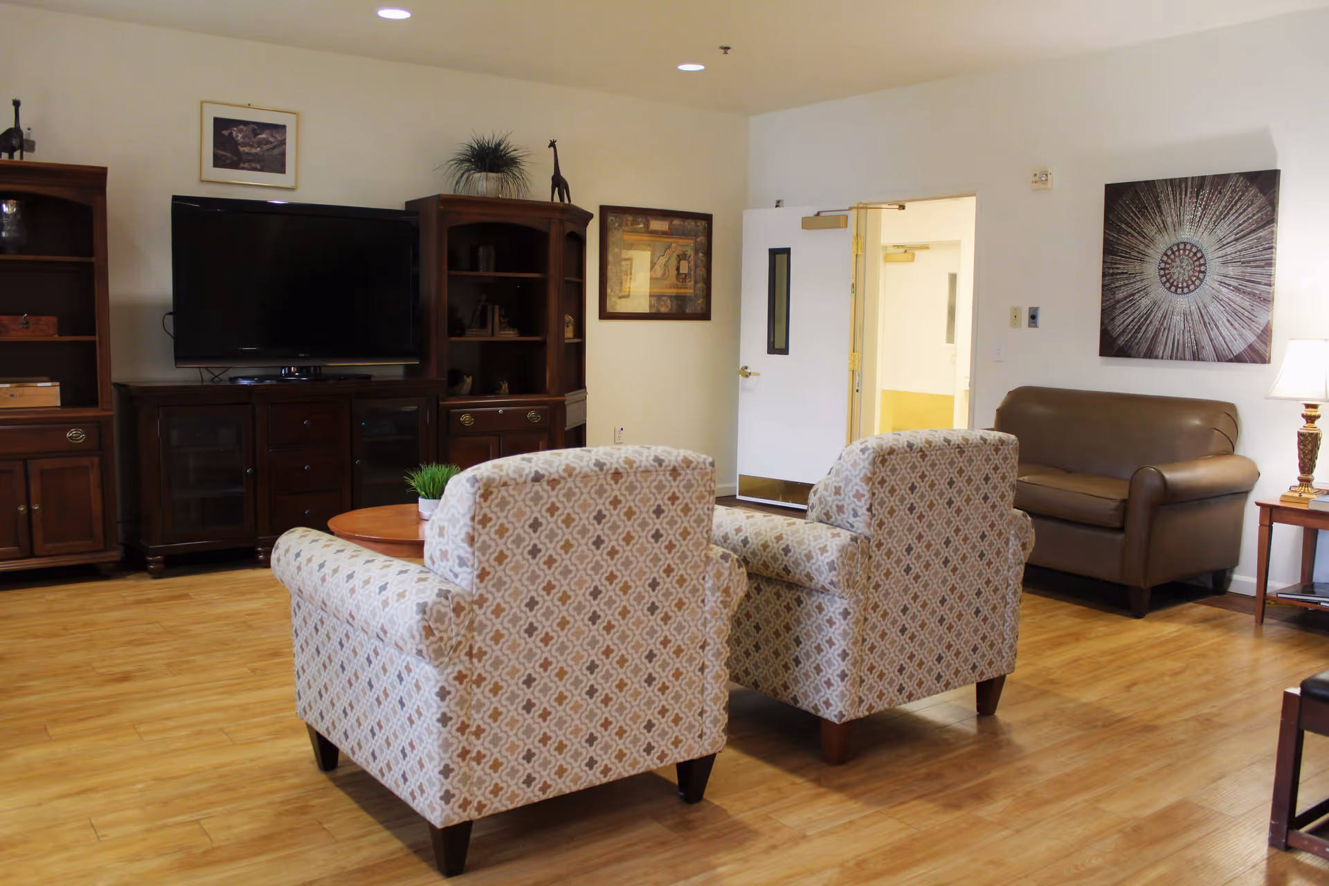 A cozy senior living facility common area with two patterned armchairs facing a wooden coffee table, a large flat-screen TV on a wooden cabinet, a brown leather loveseat, a side table with a lamp, and framed artwork on the walls. The room has wooden flooring and a white door leading to another area.