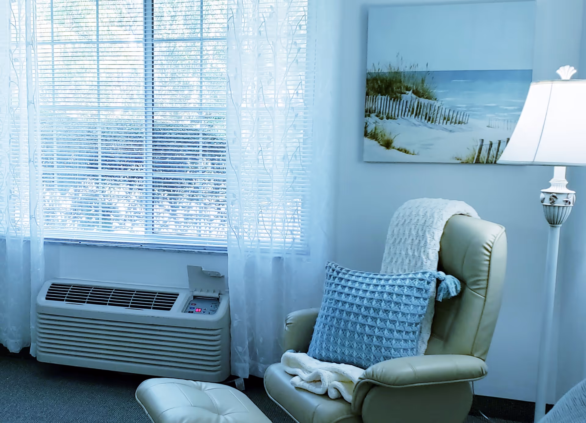 A cozy corner of a room with a light green recliner chair draped with a white knitted blanket and a textured blue pillow. Next to the chair is a matching ottoman. A floor lamp with a white shade stands nearby, and a painting of a beach scene hangs on the wall. The window behind has white blinds and sheer white curtains, with a wall-mounted air conditioning unit below it.