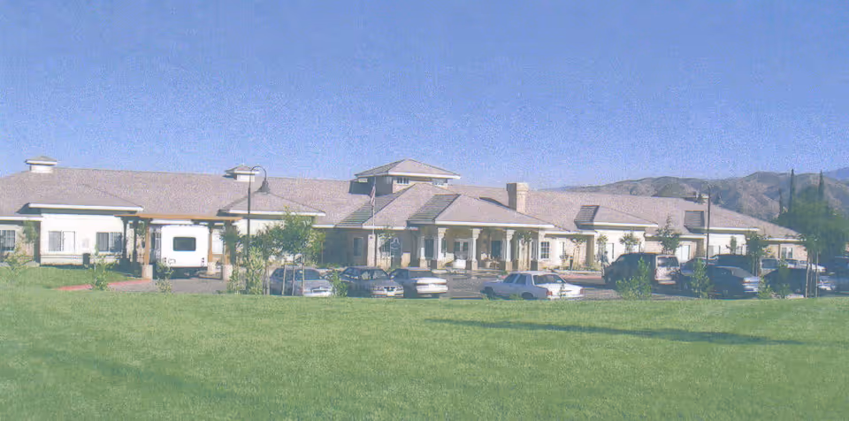 Exterior view of Wildwood Canyon Villa Assisted Living and Memory Care building with a large green lawn in the foreground, several parked cars, and mountains in the background under a clear blue sky.