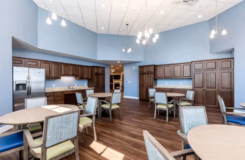 Communal dining area with round tables and chairs, a built-in kitchenette with dark wood cabinets and stainless steel refrigerator, and pendant lighting under a high ceiling.