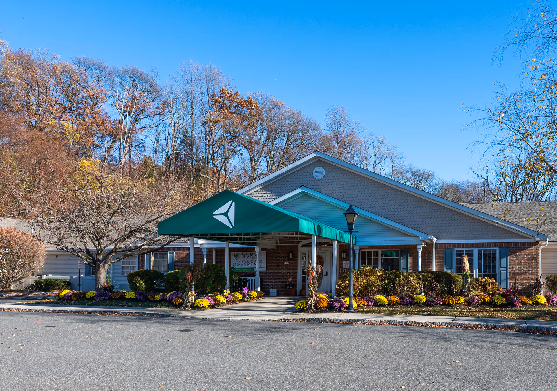 Front exterior view of Arden Courts - ProMedica Memory Care Community (Wayne) building with a green canopy entrance, surrounded by colorful flowers and trees with autumn foliage under a clear blue sky.