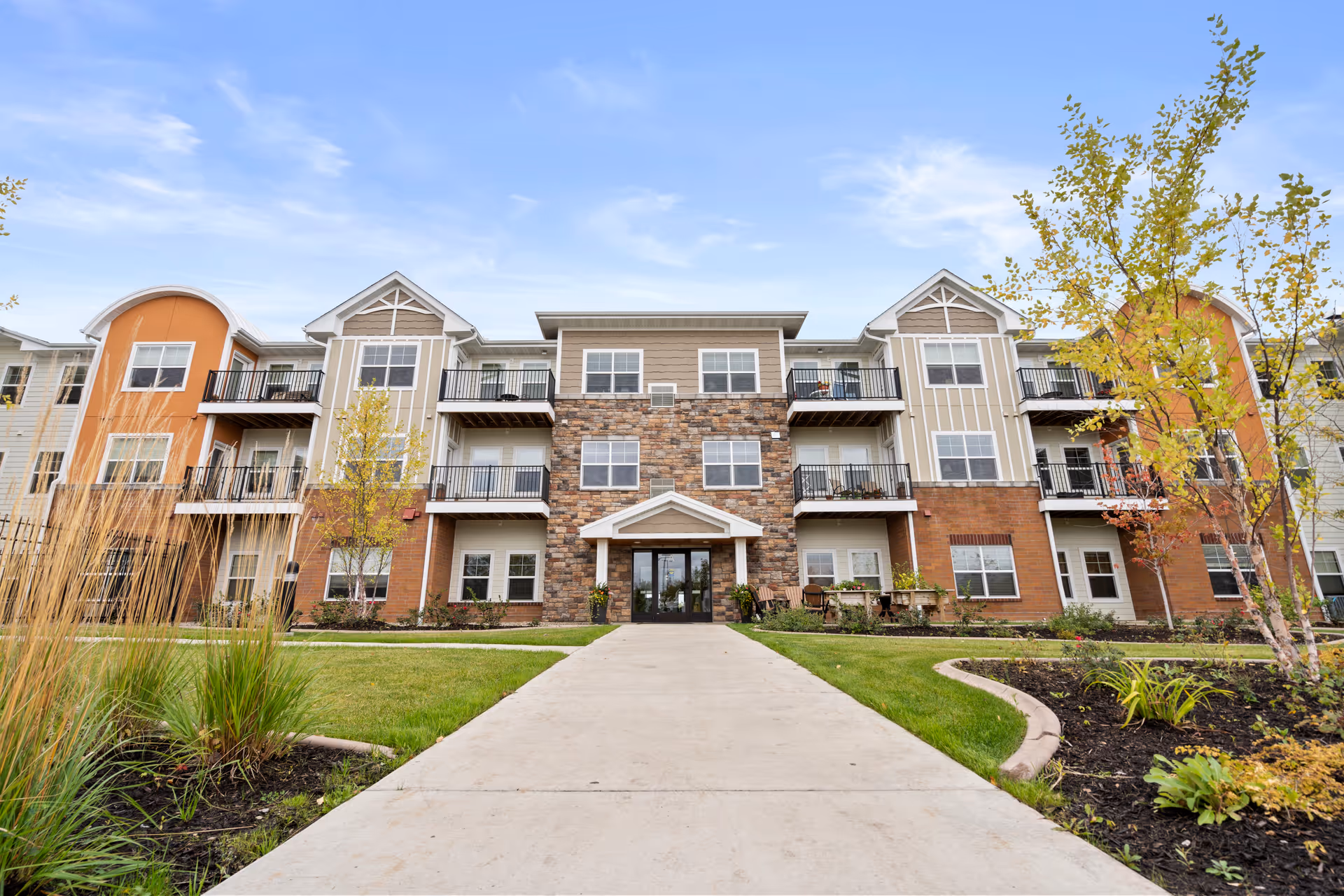 Front exterior view of a three-story senior living facility building with a stone and brick facade, balconies, and a paved walkway leading to the main entrance. The building is surrounded by landscaped grass, plants, and small trees under a partly cloudy sky.