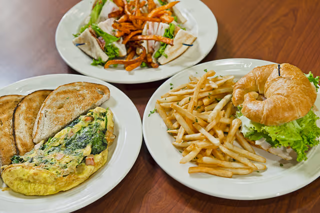 Three plates of food on a wooden table. One plate has an omelet with spinach and ham, served with three slices of toasted bread. Another plate has a croissant sandwich with lettuce and a side of French fries. The third plate has a sandwich with lettuce and turkey, served with sweet potato fries.