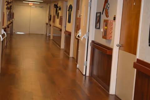 Long, well-lit interior hallway with wood floors, handrails, and multiple open doors in a nursing and rehabilitation facility.