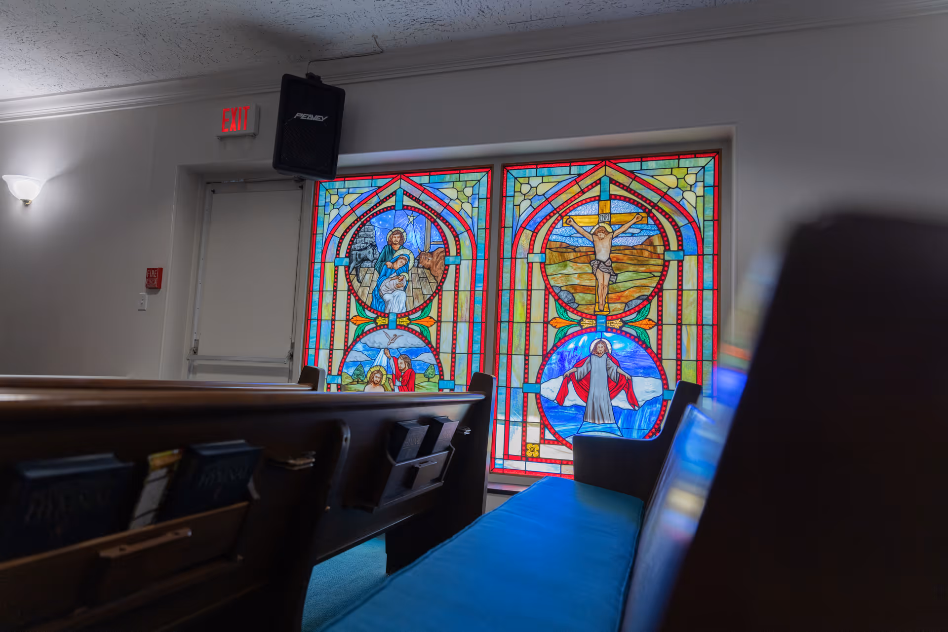 Interior view of a room with wooden pews and blue cushions facing stained glass windows depicting religious scenes, including Jesus on the cross and other biblical figures. A speaker is mounted near the ceiling above an exit door.