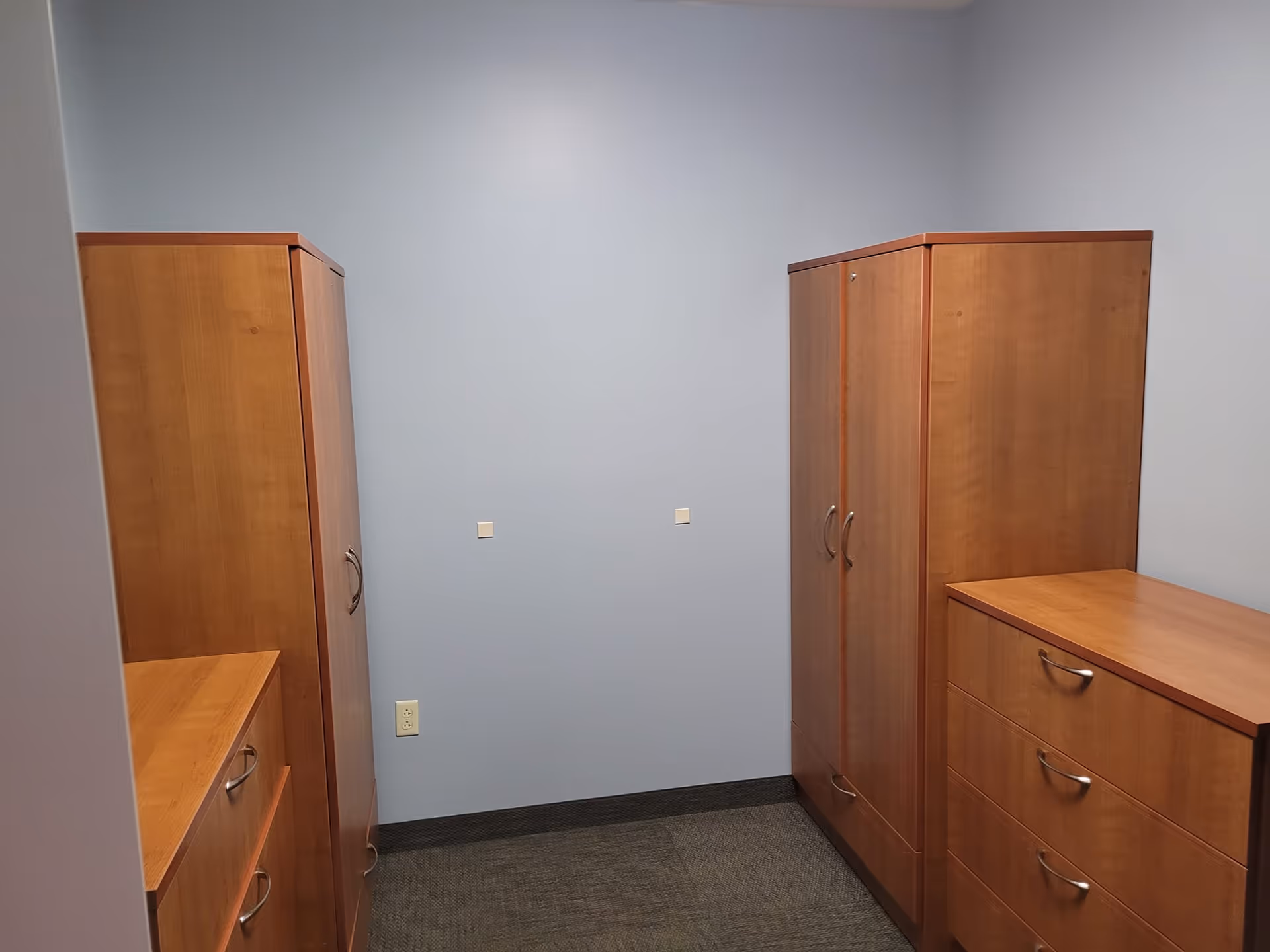 A small room with light blue walls and dark carpeted floor containing wooden storage furniture including two tall wardrobes and two dressers with drawers and silver handles.