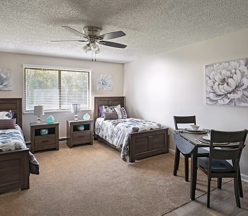 A bright bedroom with two twin beds featuring floral bedding and multiple pillows. Between the beds are two matching wooden nightstands with lamps and decorative items. A large window with blinds lets in natural light. On the right side of the room, there is a small dining table set with two chairs and place settings. The walls are decorated with floral artwork, and a ceiling fan with lights is mounted on the ceiling.