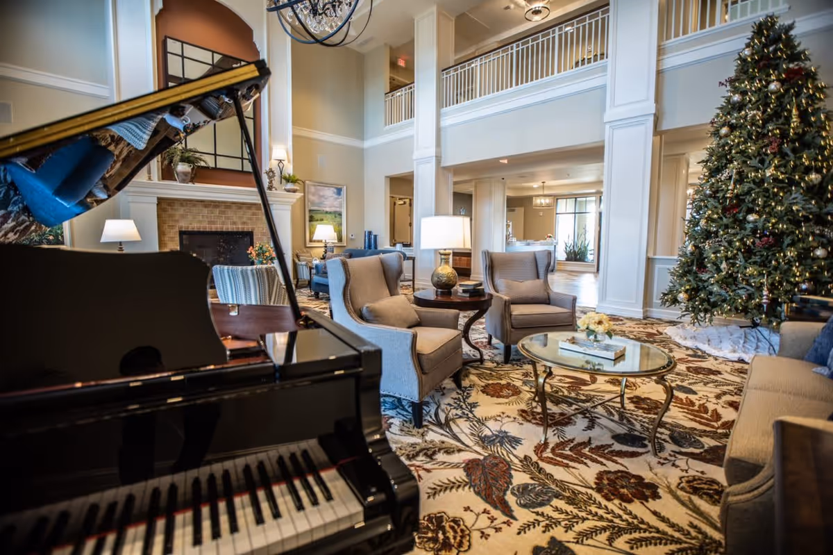 Spacious and elegantly decorated living room area with a grand piano in the foreground, comfortable armchairs, a glass coffee table, a large decorated Christmas tree, and a fireplace in the background. The room features high ceilings and a second-floor balcony overlooking the space.
