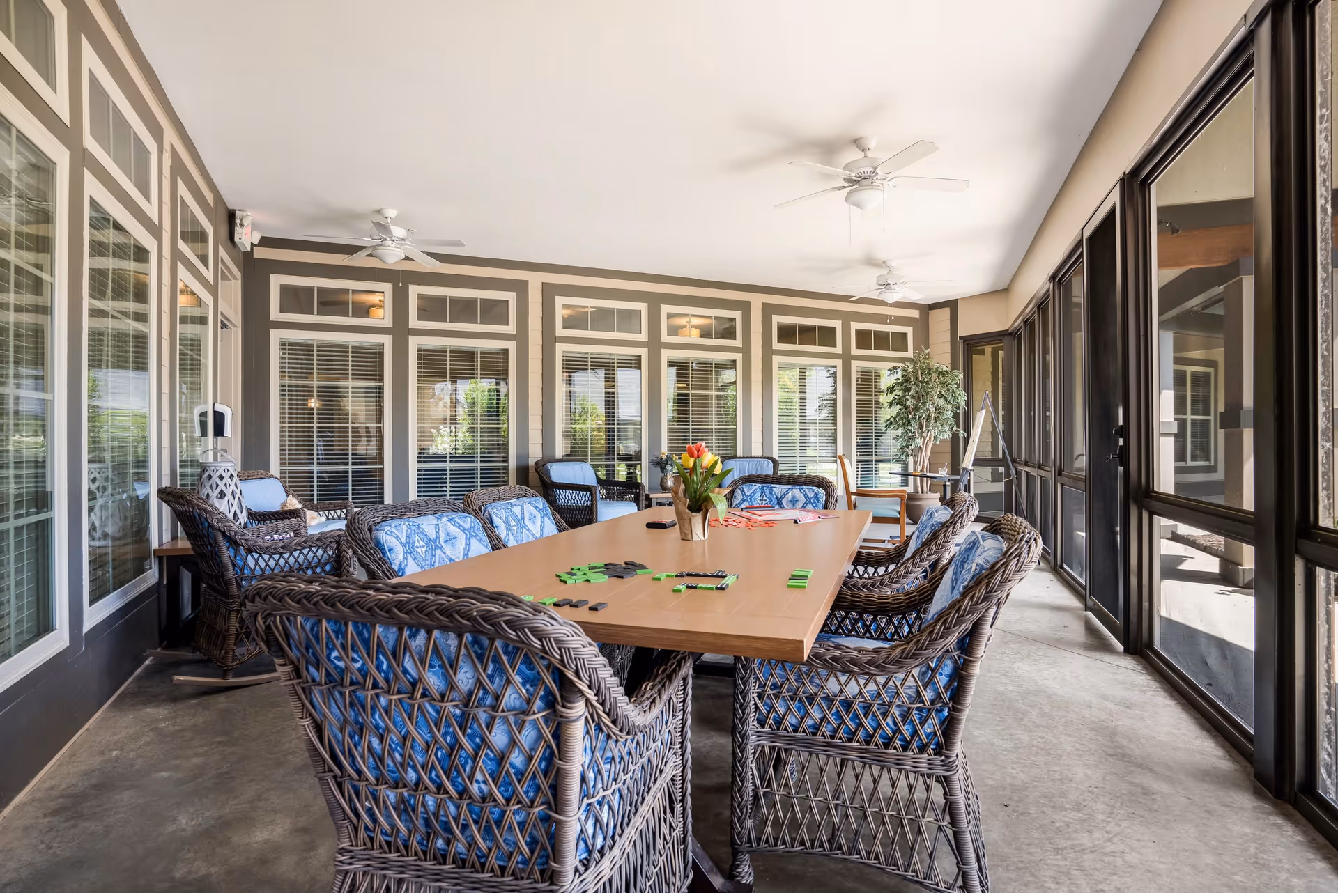 A bright and spacious indoor seating area with a large rectangular wooden table surrounded by wicker chairs with blue patterned cushions. The table has a vase with flowers and some game pieces on it. The room features large windows with white blinds, ceiling fans, and a potted plant in the corner. The floor is concrete and the walls are painted in neutral tones with white trim.