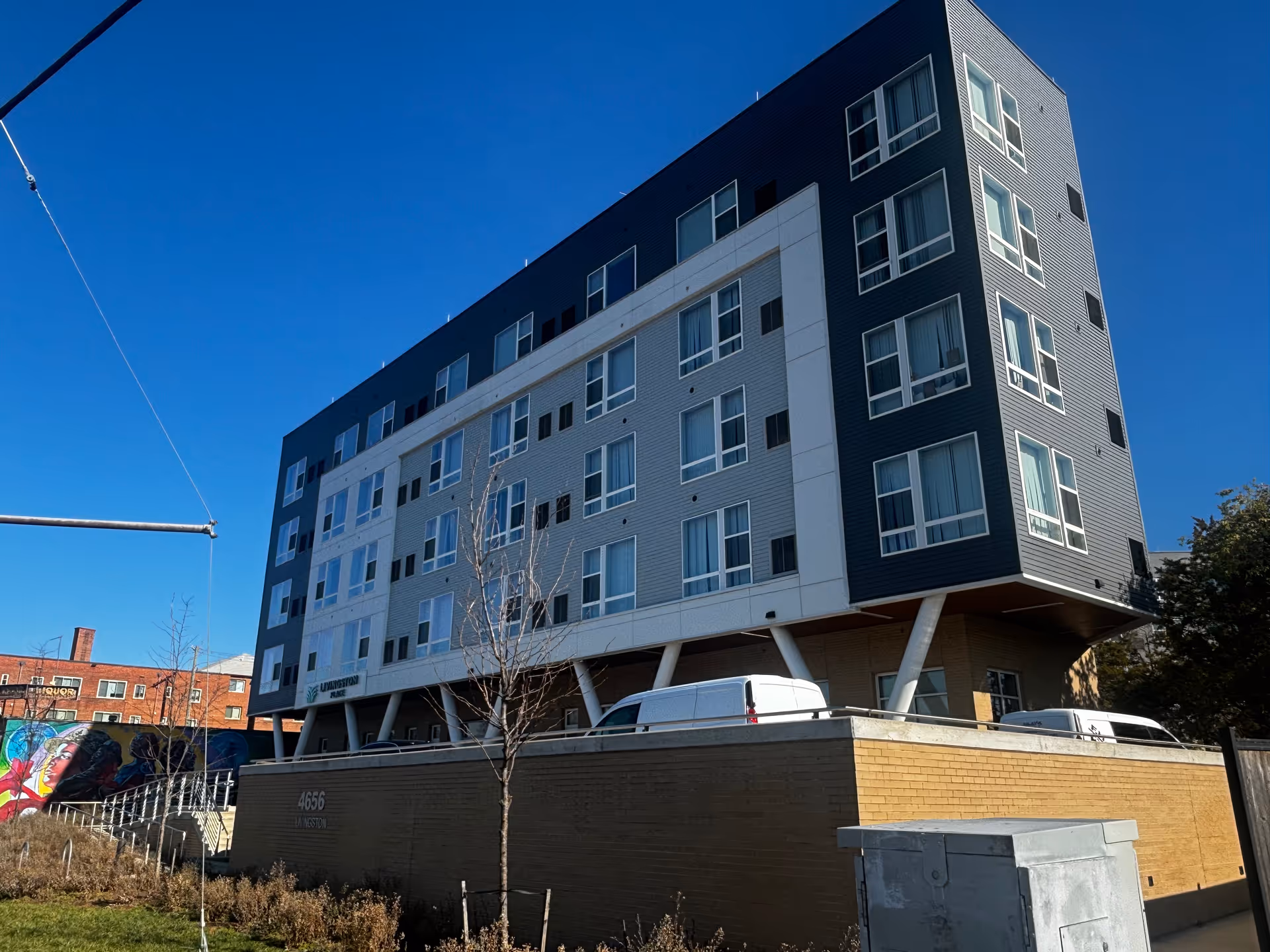Exterior view of a modern multi-story building with numerous windows under a clear blue sky. The building has a sign that reads 'Livingston Place' and is elevated on angled supports above a brick base. There are some vehicles parked near the building and a colorful mural visible on a wall to the left.