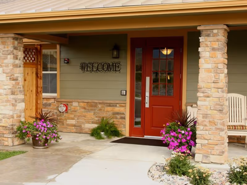 Front entrance of a building with a red door, stone pillars, and a welcome sign on the wall. There are flower pots with purple flowers on either side of the door and a bench to the right.