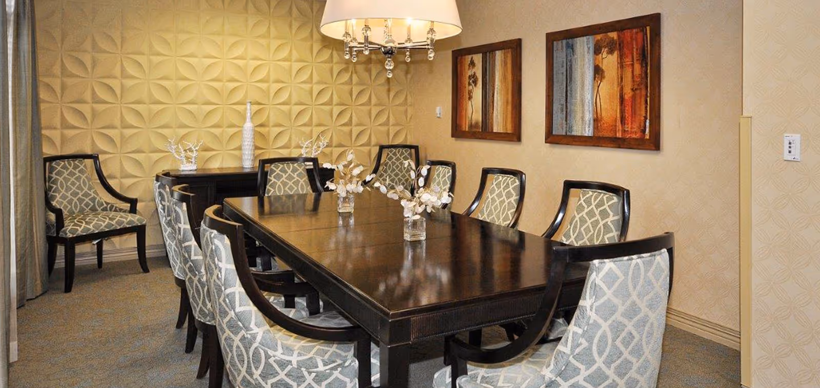 A formal dining room with a long dark wooden table surrounded by eight upholstered chairs with a geometric pattern. The room features a textured yellow accent wall, two framed abstract paintings on a beige wall, a chandelier with hanging crystals, and decorative vases on a sideboard.